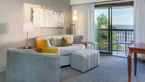A modern hotel room with a grey sofa, ottoman, and a small table. A large window offers a view of the outdoors.