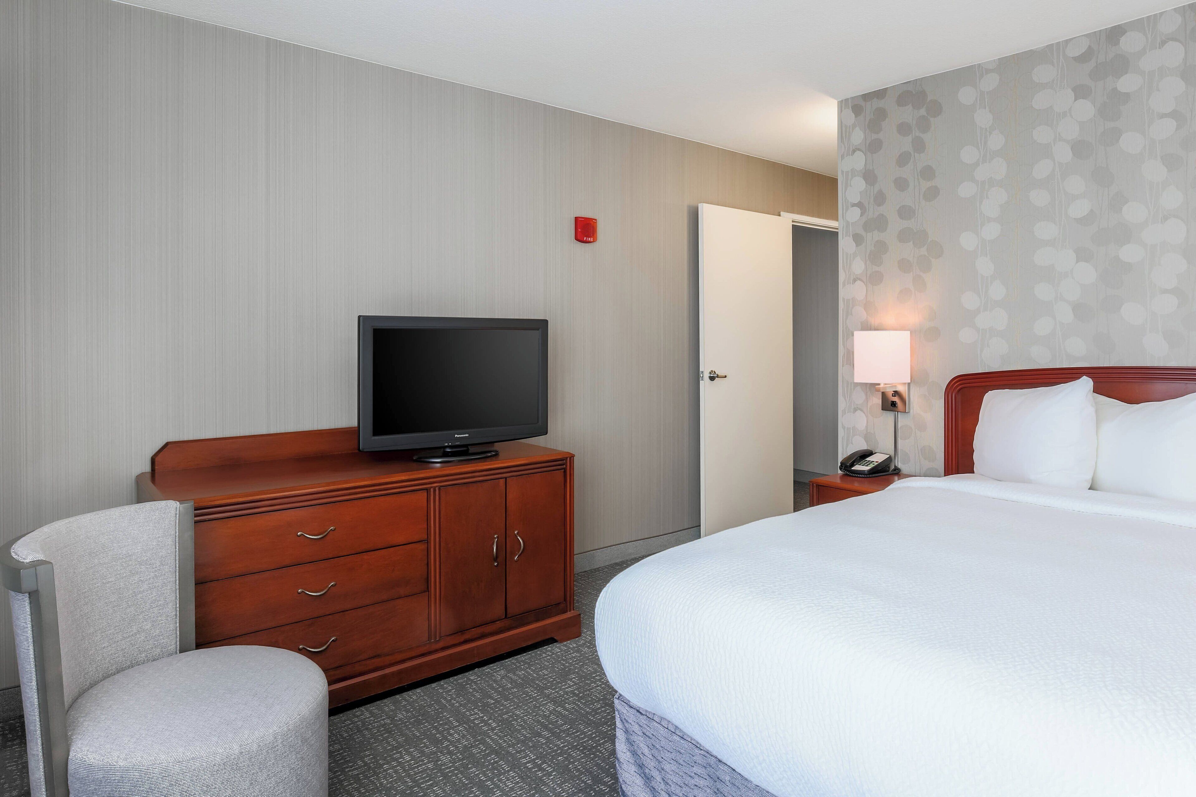A hotel room with a bed, a television on a wooden dresser, a chair, a nightstand with a lamp, and a door.
