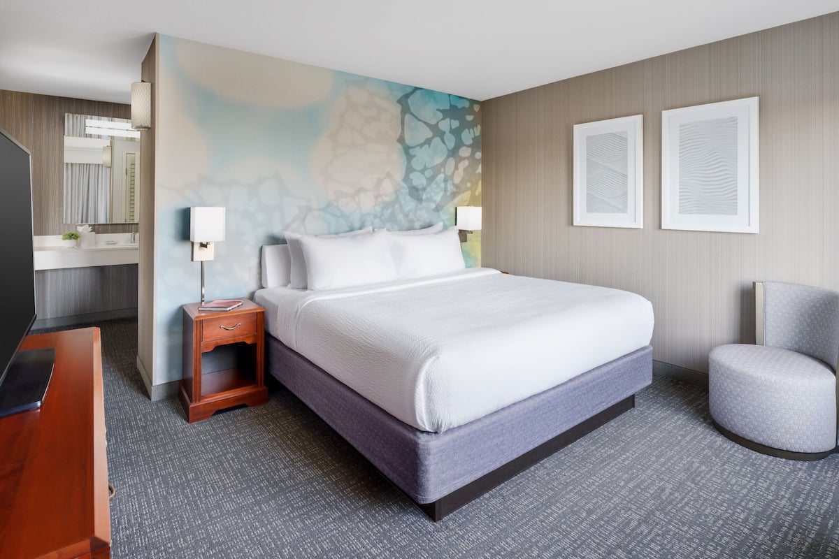 A hotel room with a large bed, a bedside table, a chair, a desk, and a mirror.