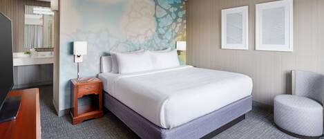 A hotel room with a large bed, a bedside table, a chair, a desk, and a mirror.