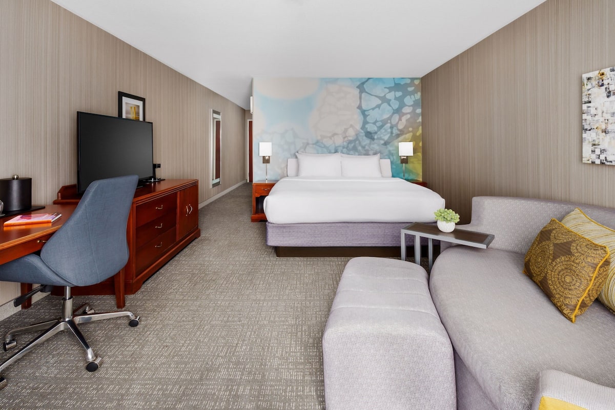 A hotel room with a bed, a sofa, a desk with a chair, a TV, and a painting on the wall.
