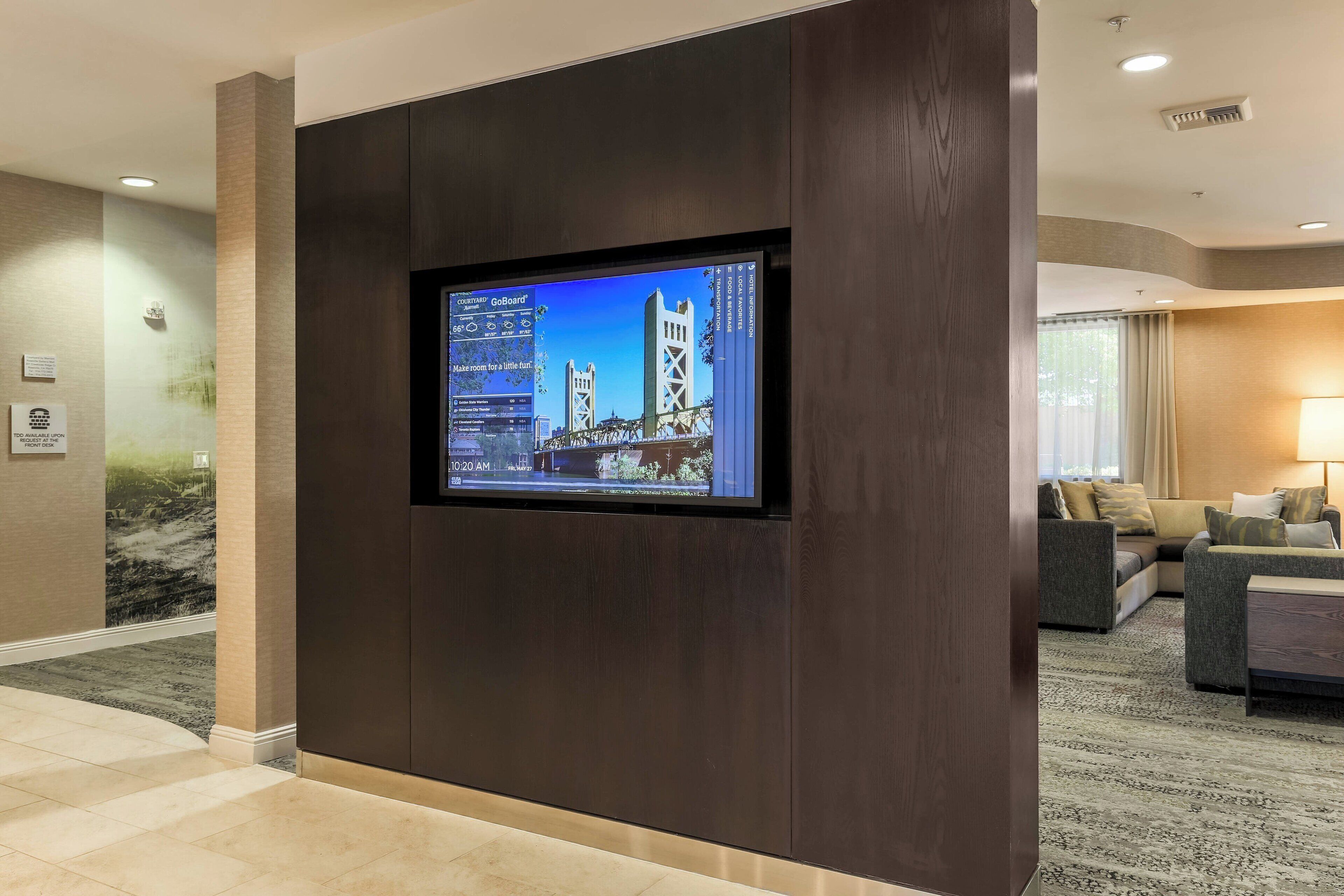 Um saguão de hotel com uma grande tela digital exibindo uma vista urbana, um balcão de recepção e uma área de estar com sofás.