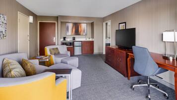 A hotel room with a sofa, a desk with a chair, a television, a kitchen area, and a large window.