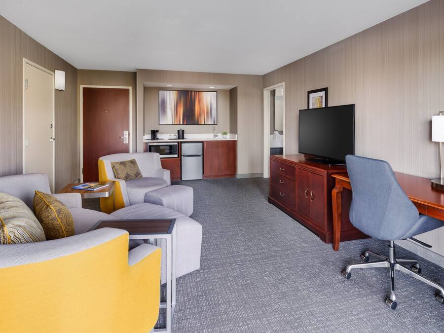 A hotel room with a sofa, a desk with a chair, a television, a kitchen area, and a large window.