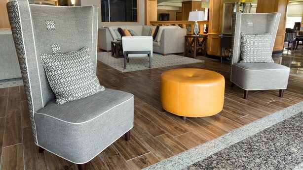A modern lobby with grey upholstered armchairs, a round wooden ottoman, and a patterned rug. - Drury Inn & Suites Lafayette, LA (Lafayette)
