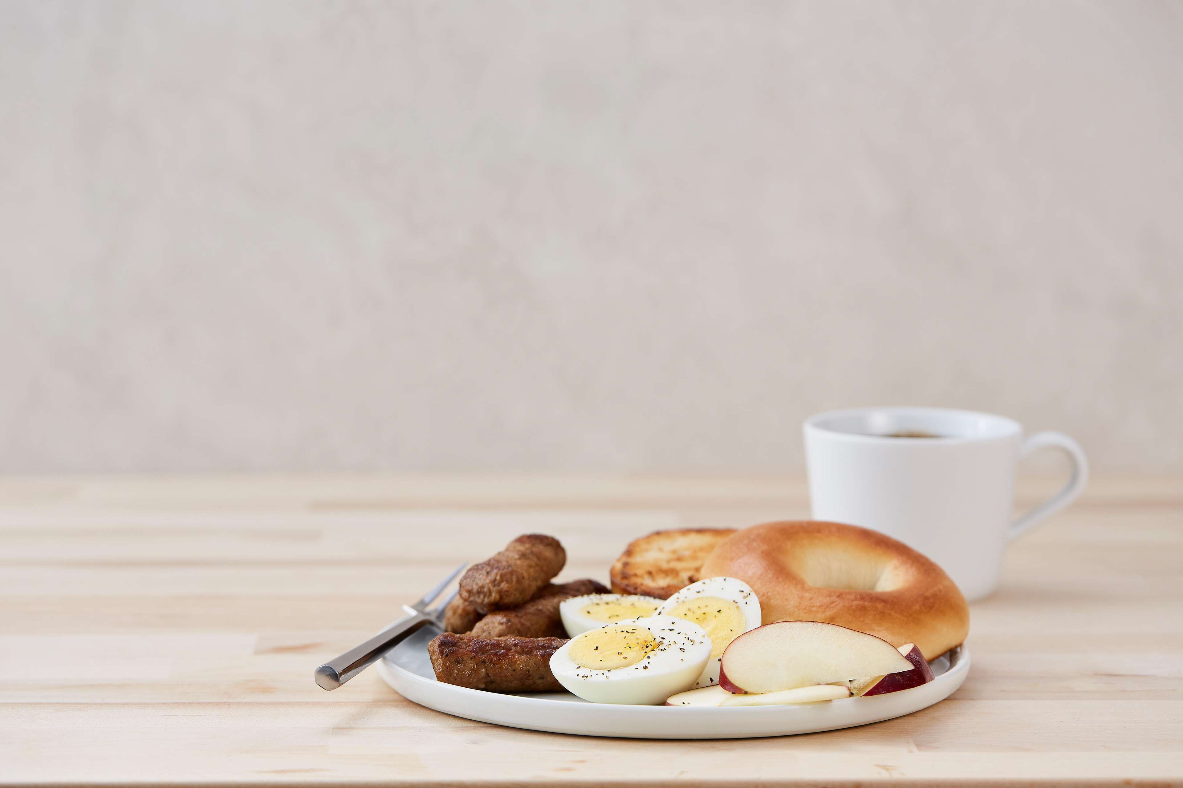 A plate with a bagel, sliced hard-boiled eggs, sausage links, and apple slices, accompanied by a white mug.