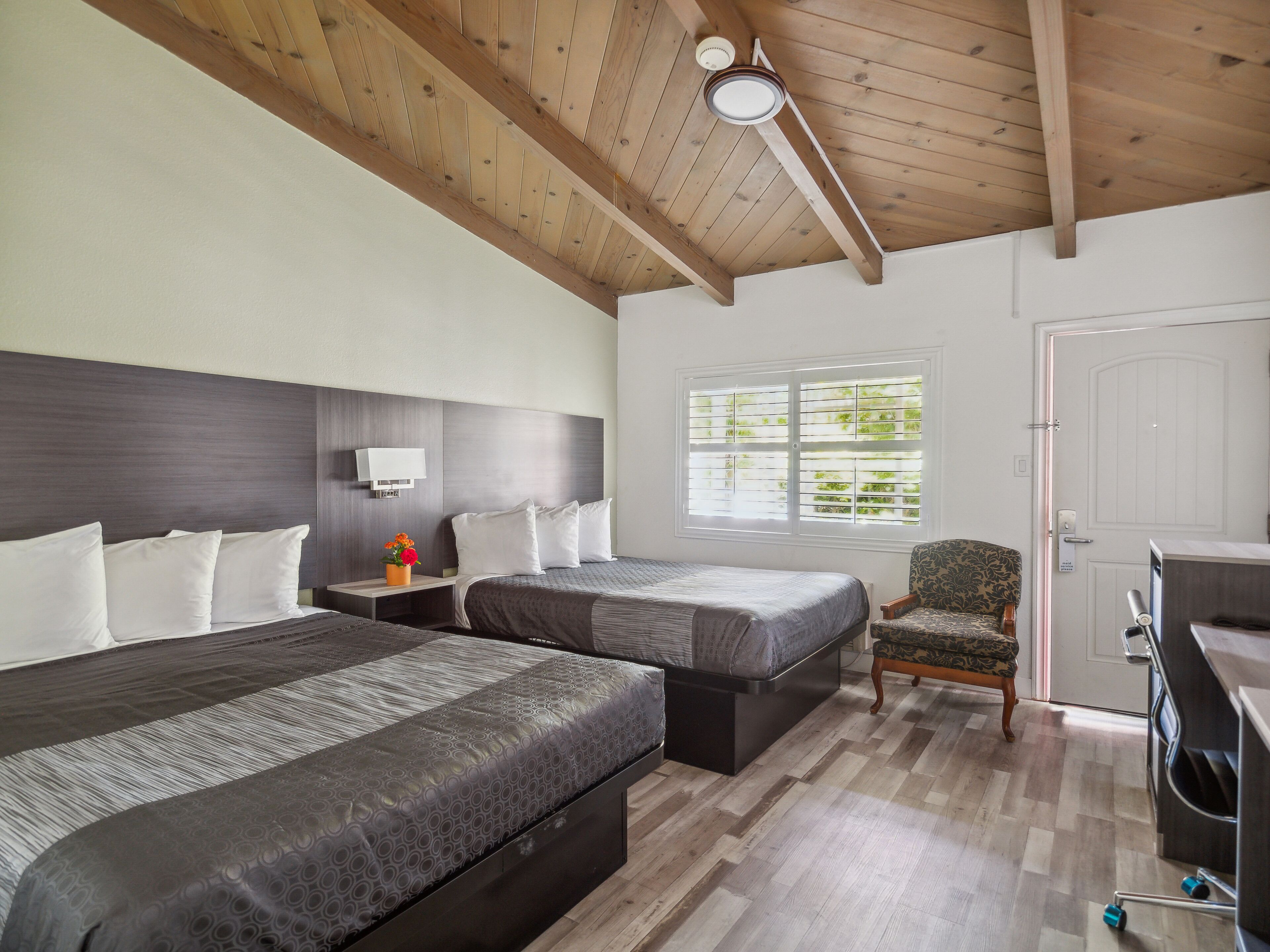 A hotel room with two beds, a wooden ceiling, a window with blinds, a chair, and a desk.