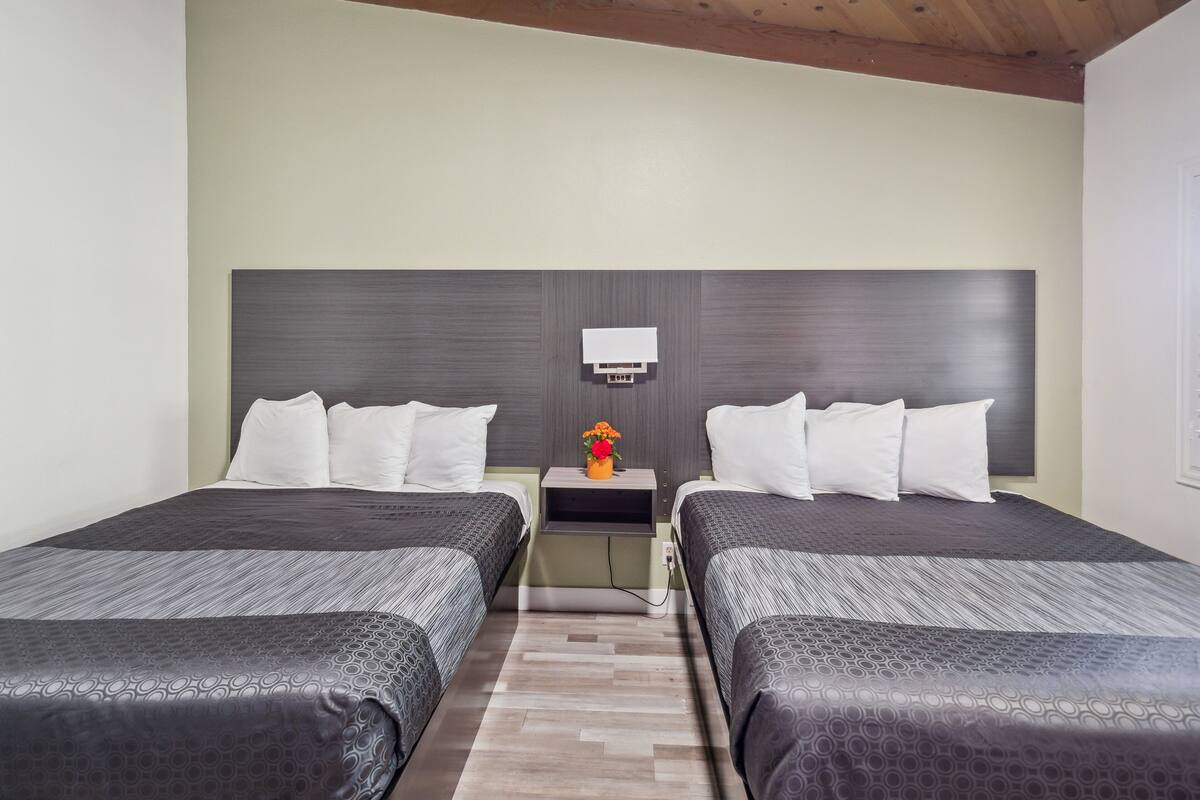 A hotel room with two beds, a nightstand, a wall-mounted lamp, and a small table with a vase of flowers.