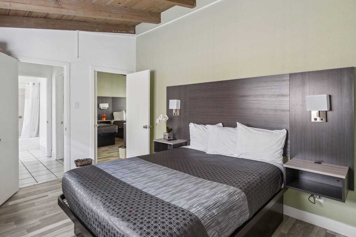 A modern hotel room with a large bed, wooden ceiling beams, a small bedside table with a lamp, and a view into the bathroom.
