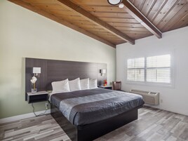 Un dormitorio con cama, mesitas de noche, lámpara, ventana con persianas y techo de madera.