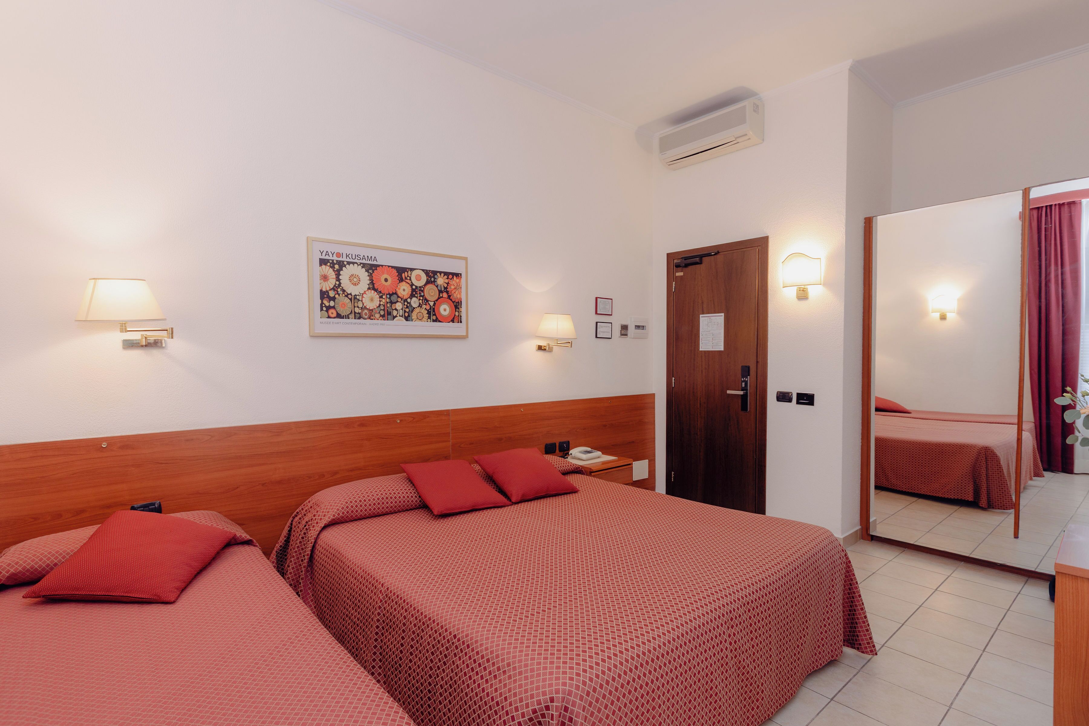 Une chambre d’hôtel avec deux lits, une tête de lit en bois, un climatiseur fixé au mur, un tableau encadré et un miroir qui reflète la pièce.