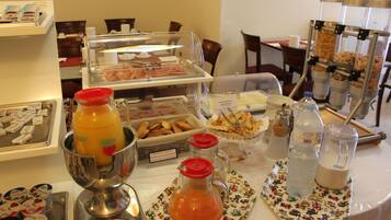 Buffet com diversos alimentos e bebidas, incluindo dispensador de ponche de frutas, estação de bebidas quentes e variedade de doces e sanduíches.