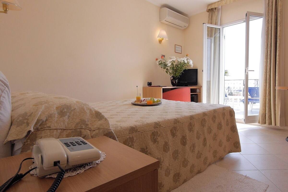 Habitación de hotel con cama, mesita de noche con teléfono, una pequeña mesa con un cuenco, televisión y balcón con una silla.