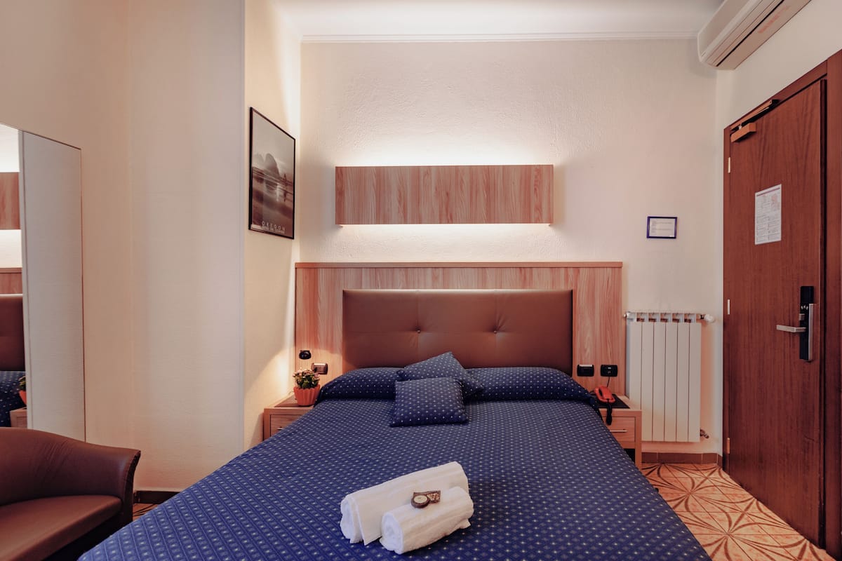 Una habitación de hotel con una cama grande, cabecera de madera, mesita de noche con lámpara y un radiador.