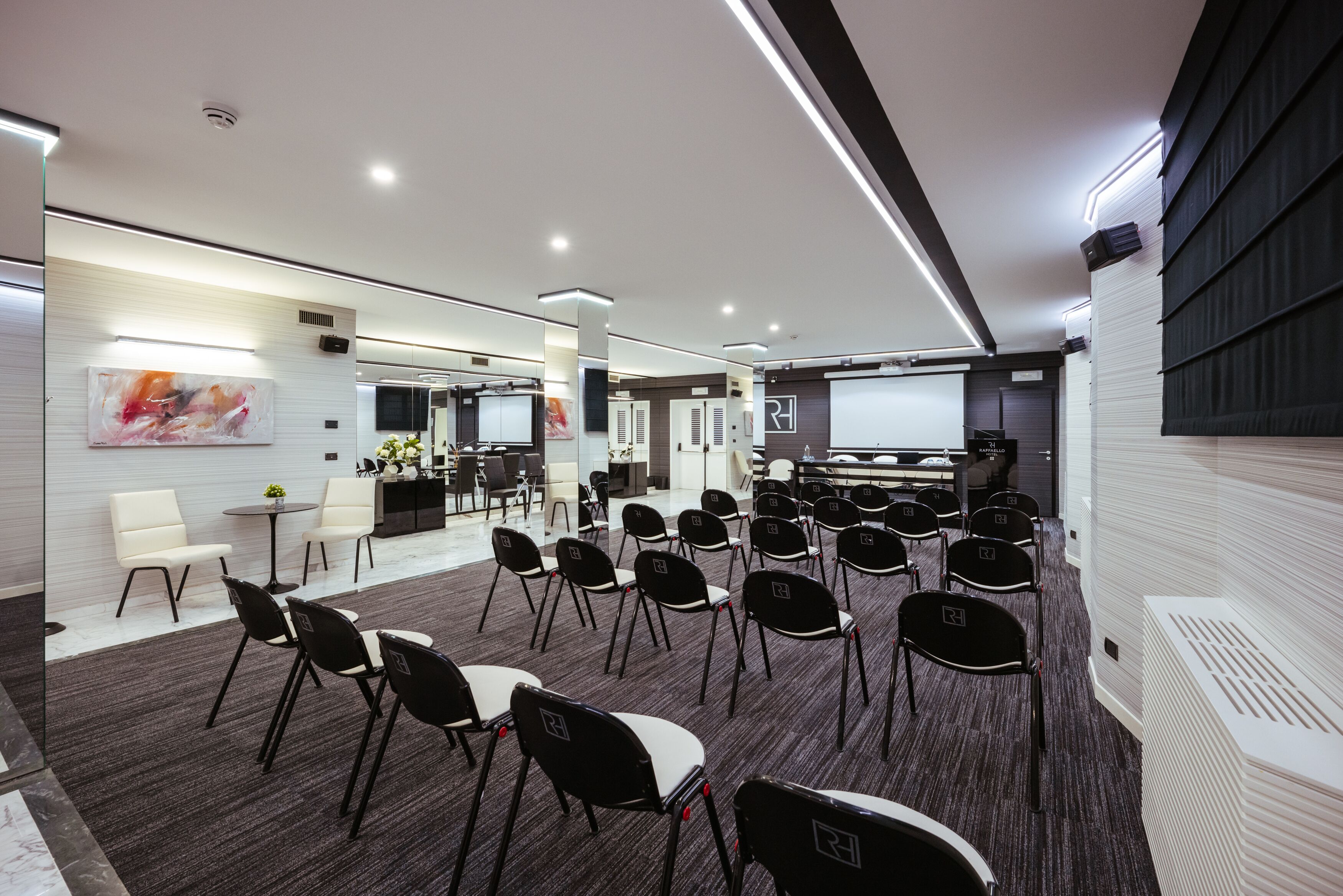 Una sala de conferencias moderna con filas de sillas negras, alfombra blanca y una pantalla grande al frente.