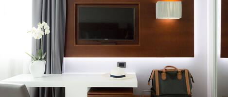 A modern hotel room with a flat-screen TV mounted on a wooden panel, a desk with a laptop, a vase with white flowers, and a handbag on the desk.