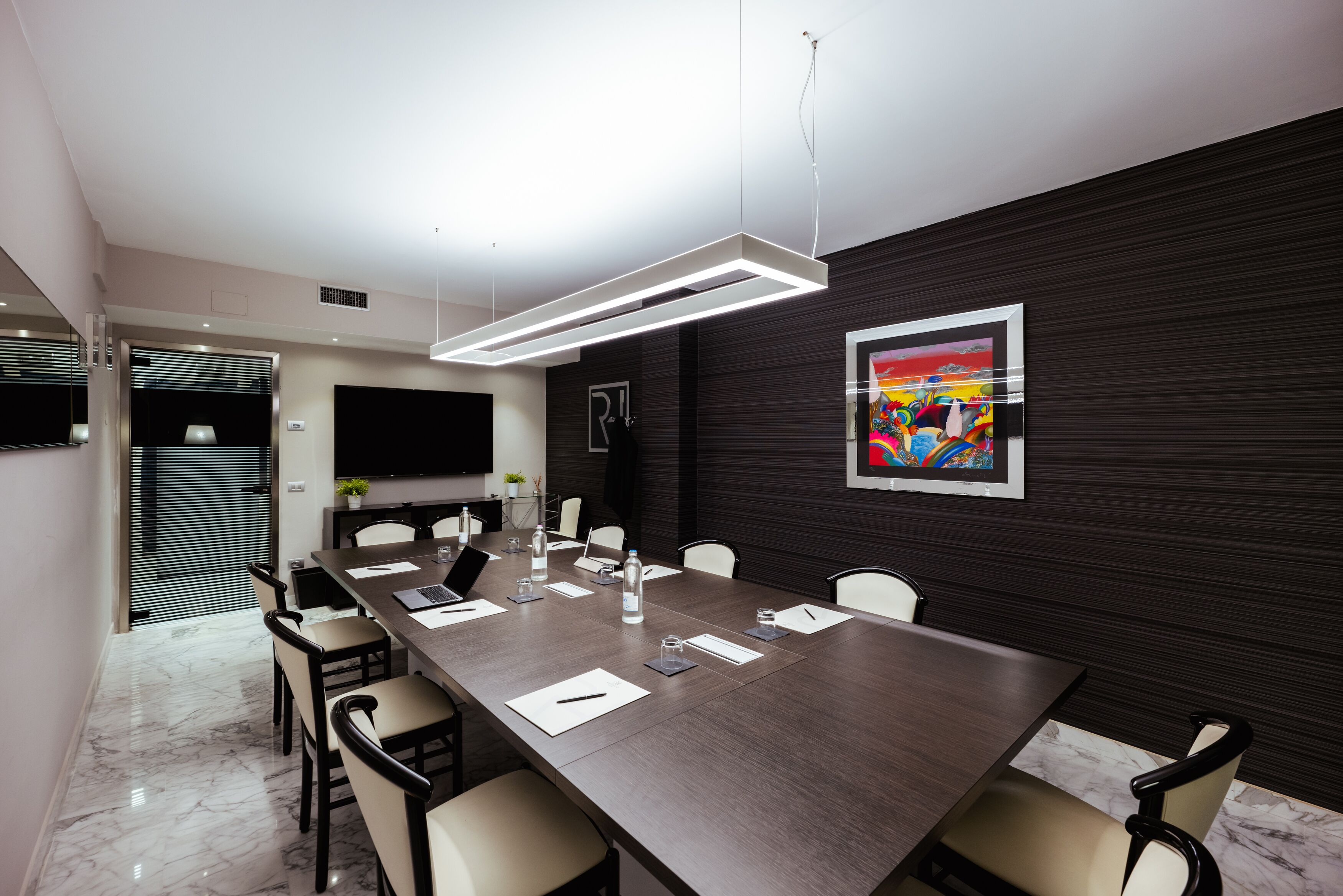 A modern conference room with a long table, chairs, a flat-screen TV, and a painting on the wall.