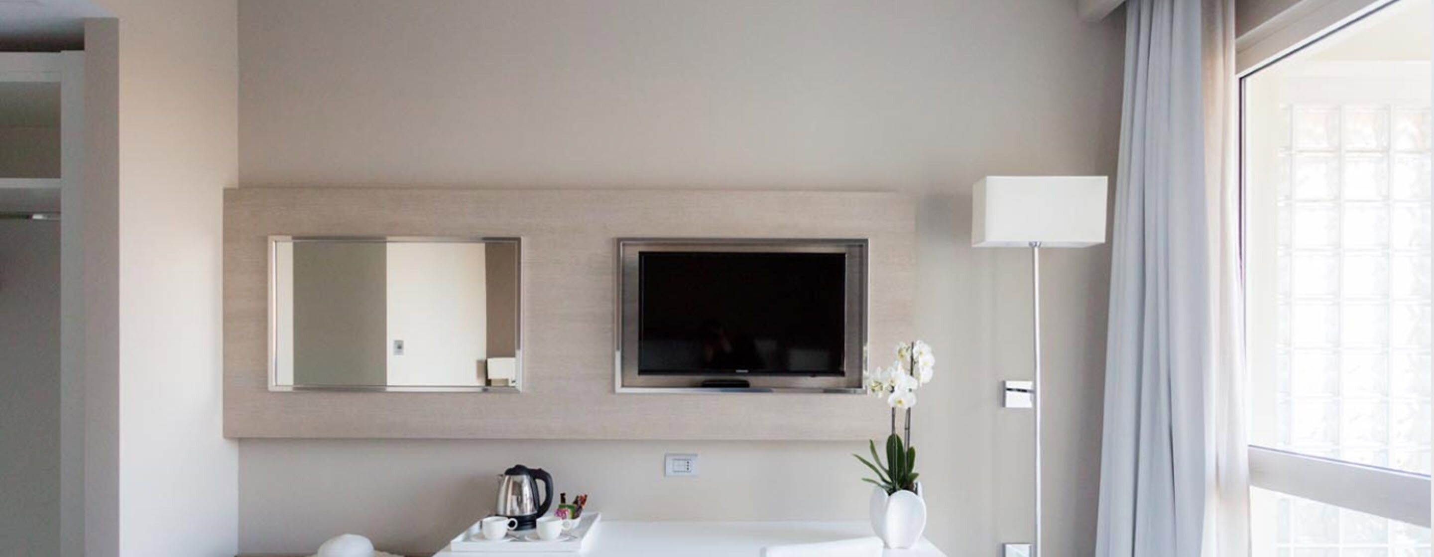 Un baño moderno con un espejo grande, un televisor de pantalla plana, un lavabo blanco y un florero con flores.