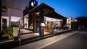 A modern restaurant with an outdoor seating area, a glass facade, and a well-lit entrance.