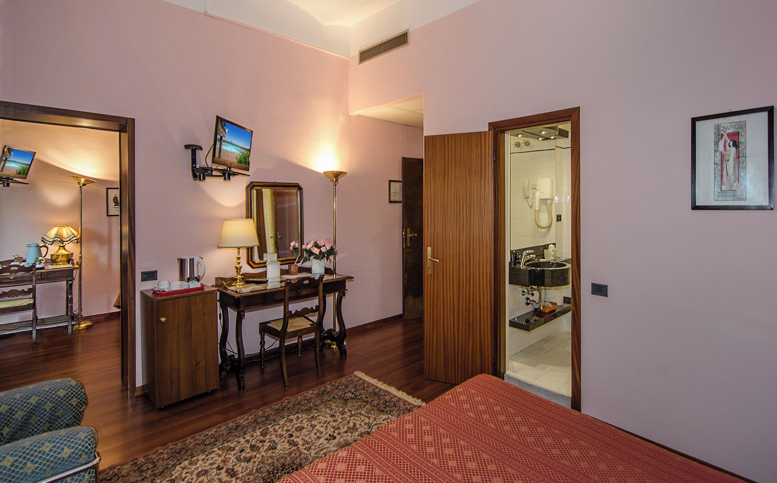 A hotel room with a bed, a desk with a chair, a mirror, a television, a bathroom with a sink and a toilet, and a door.