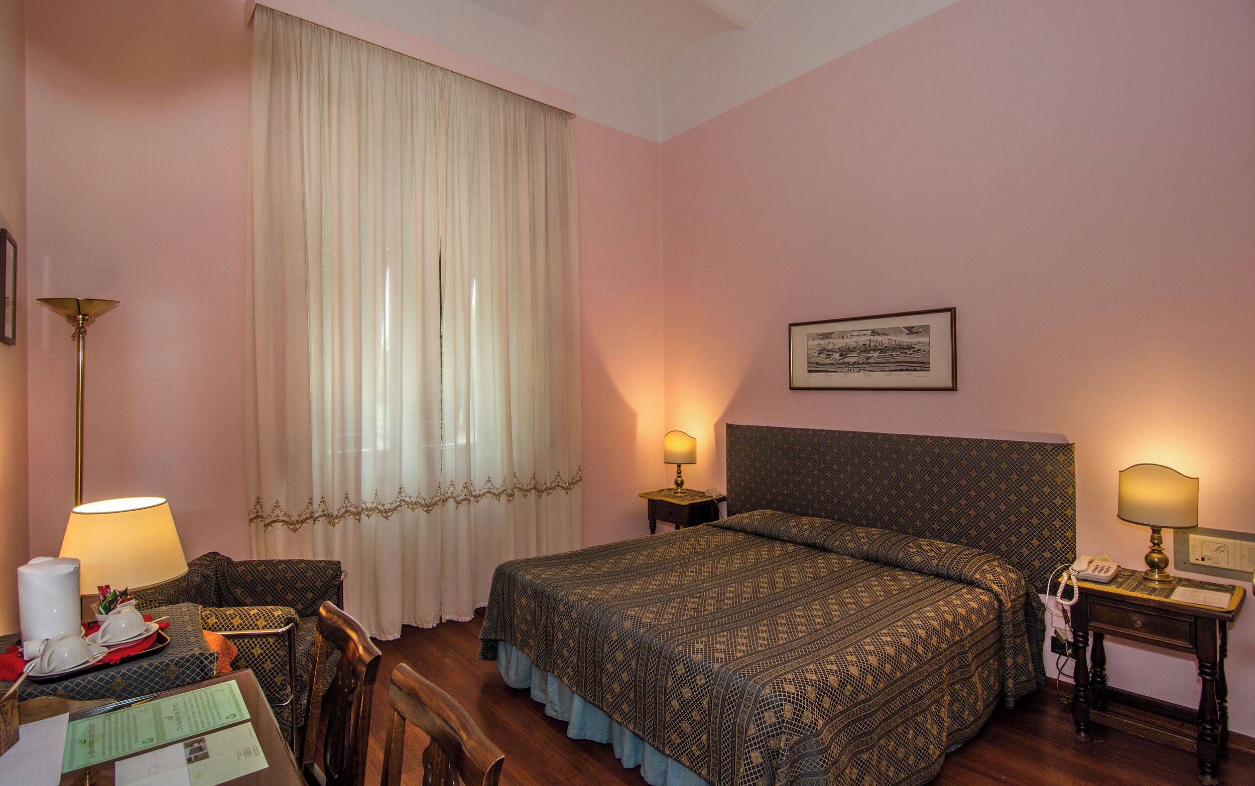 A hotel room with a bed, two bedside tables, a desk with a chair, a lamp, and a framed picture on the wall.