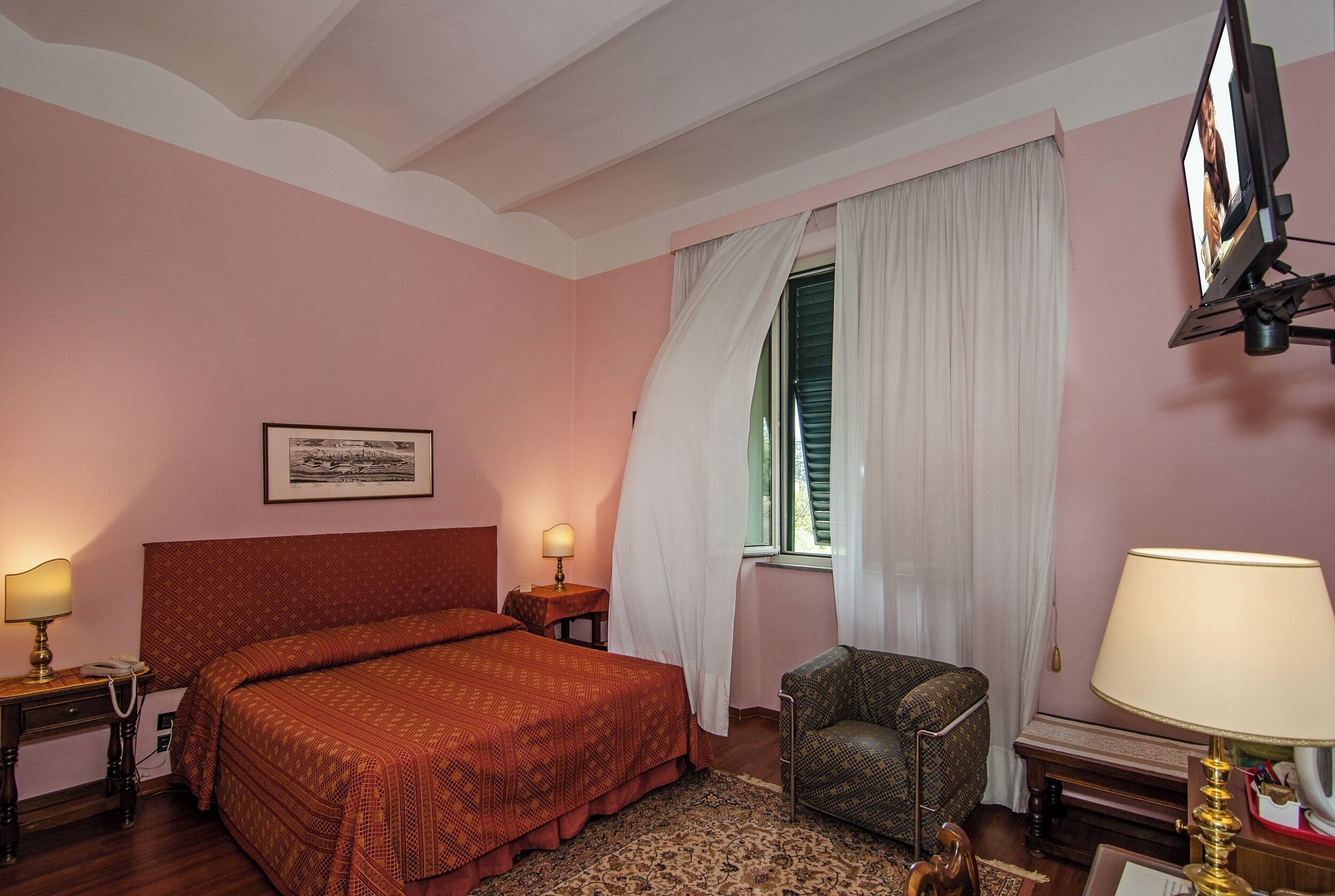 Una camera d'albergo con un letto, una sedia, una scrivania con una lampada, un quadro appeso al muro e una finestra con tende.