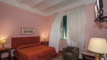 Una camera d'albergo con un letto, una sedia, una scrivania con una lampada, un quadro appeso al muro e una finestra con tende.