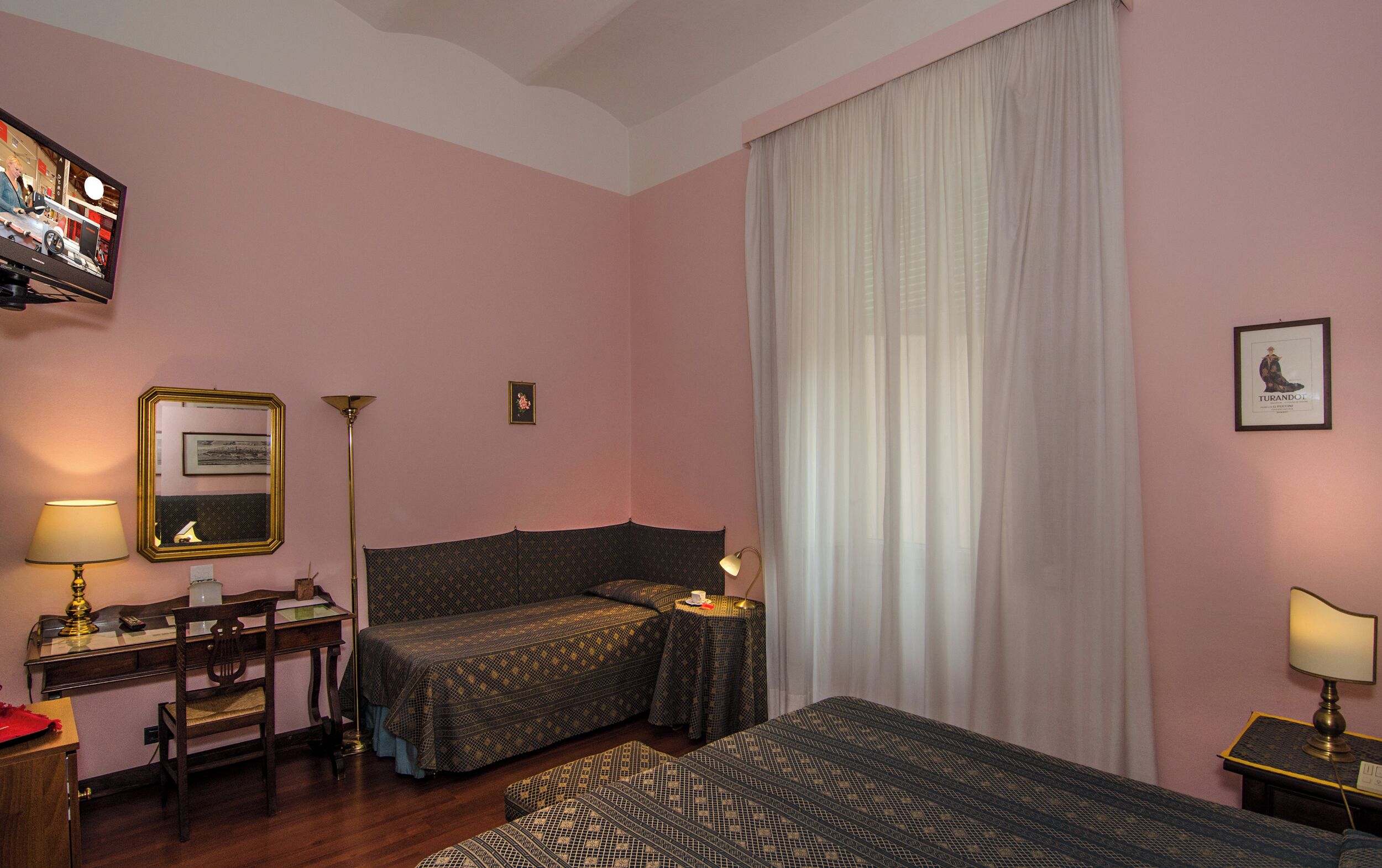 A hotel room with a bed, a desk, a chair, a lamp, a television, and a large window with curtains.