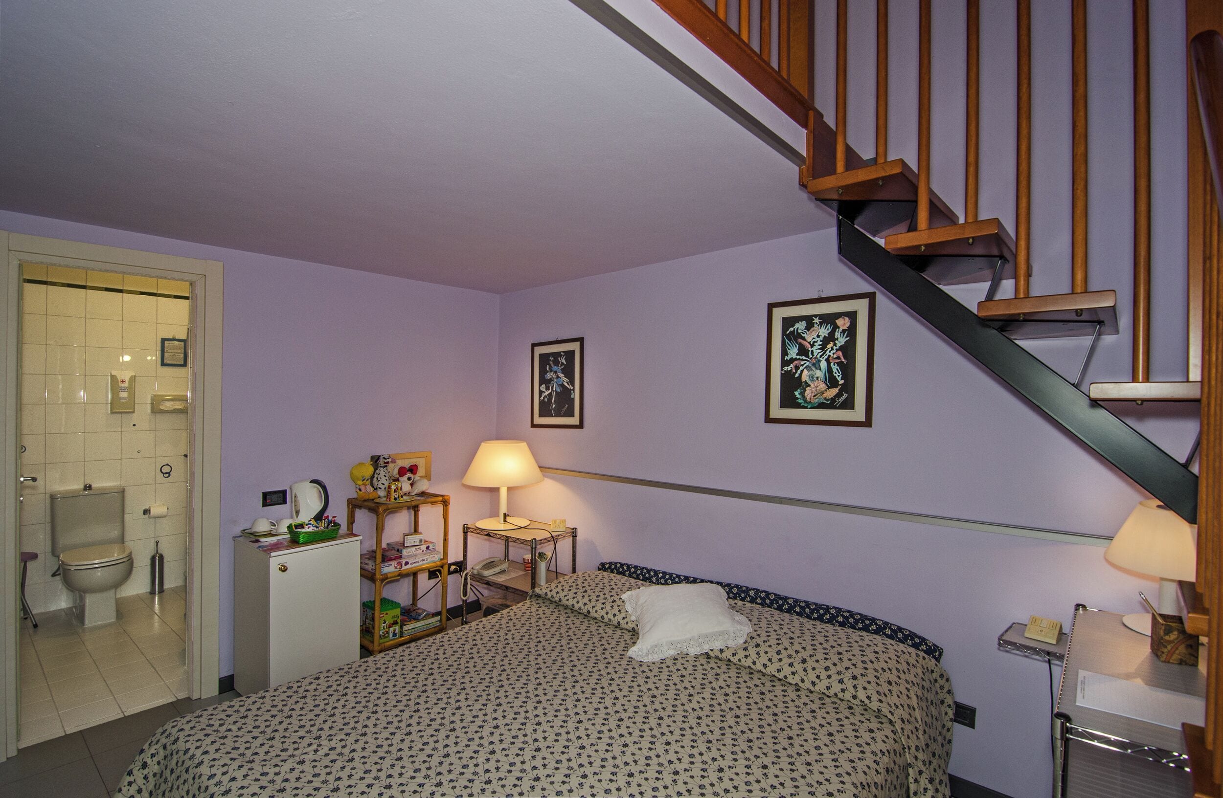 A hotel room with a bed, a desk, a lamp, a small refrigerator, a bathroom, and a wooden staircase.