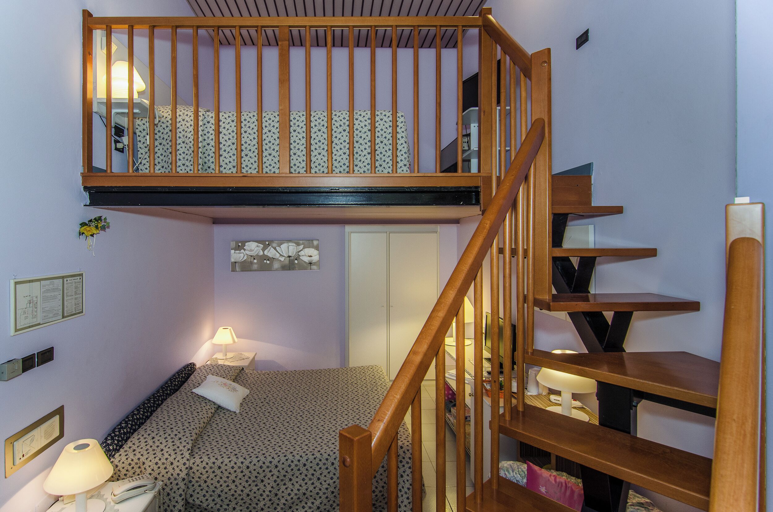 Una habitación con una escalera de madera, una cama con ropa de cama floral, una lámpara de noche y una mesita con lámpara.