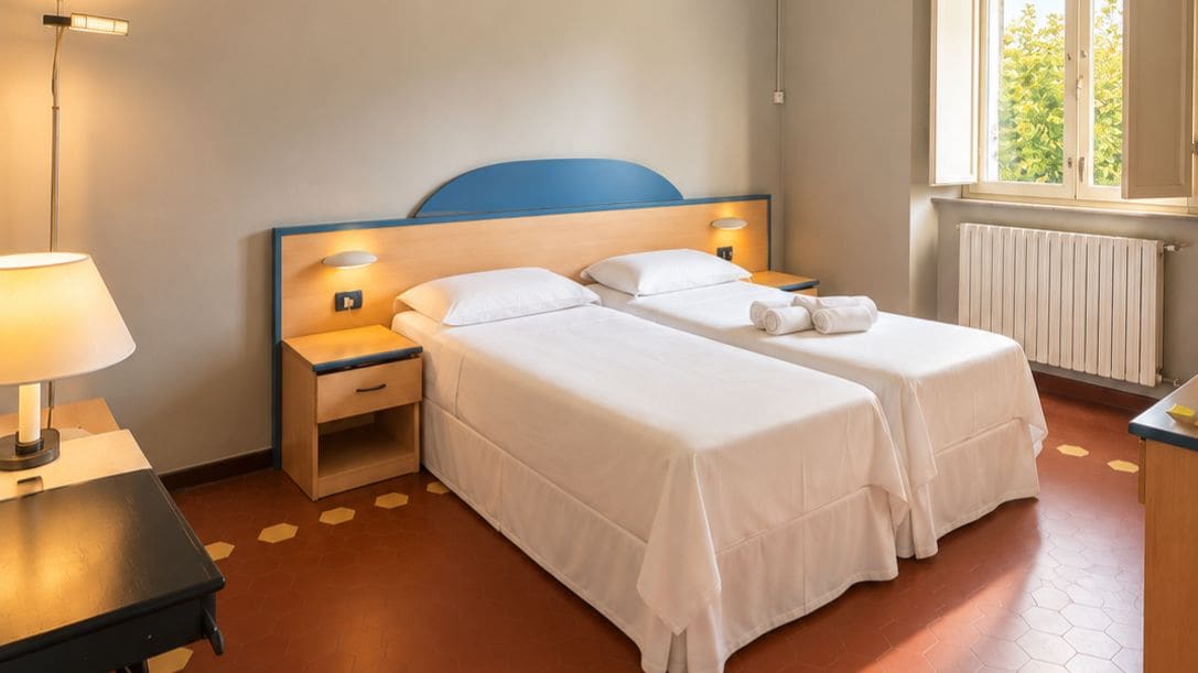 Habitación de hotel con dos camas, una mesita de noche, una lámpara, un ventanal con vistas a la vegetación y un radiador.