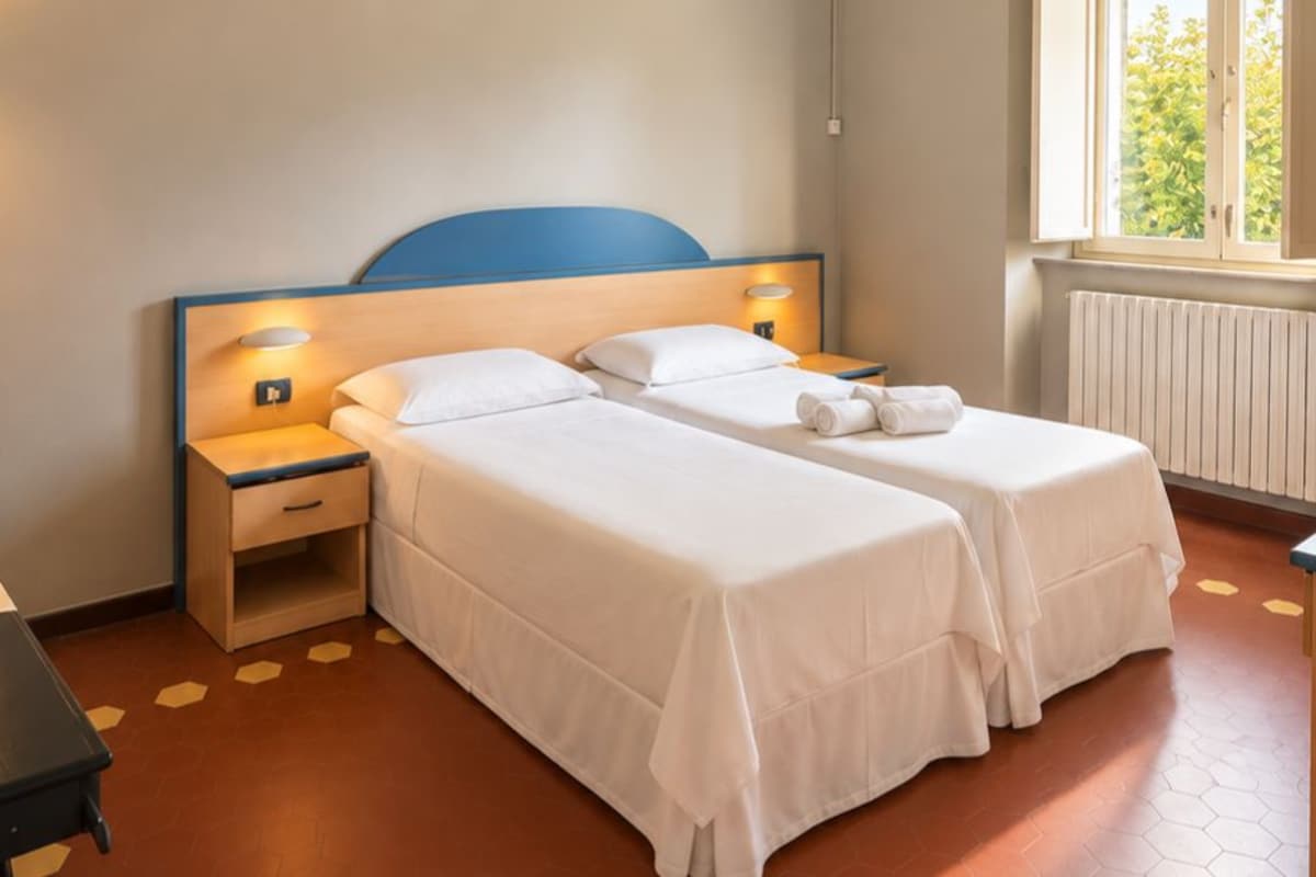 Hotellihuone, jossa on kaksi sänkyä, yöpöytä, lamppu, ikkuna, josta on näkymä vehreään maisemaan, ja patteri.