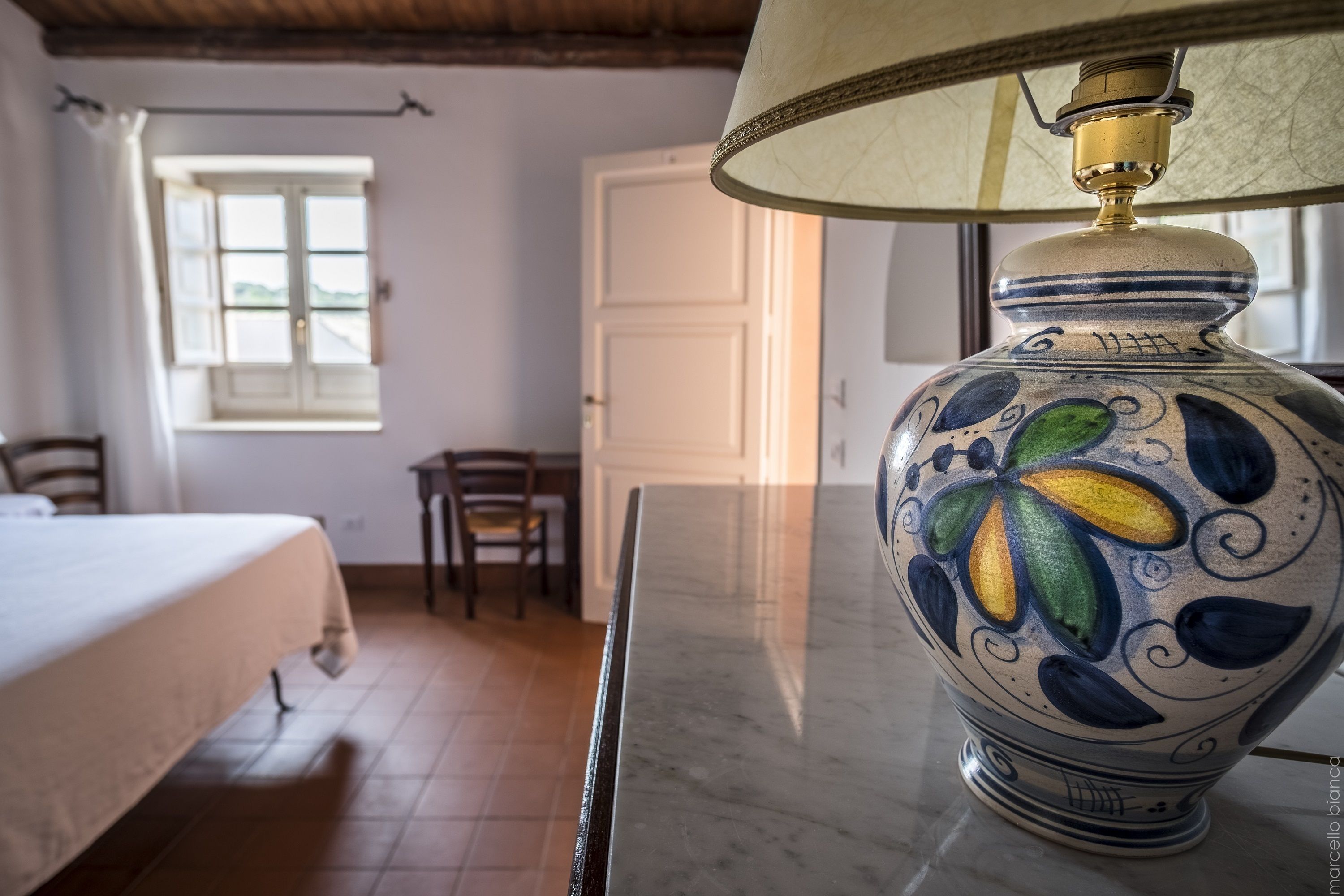 Una habitación con una cama, una mesa con una lámpara decorativa, una ventana y una puerta.