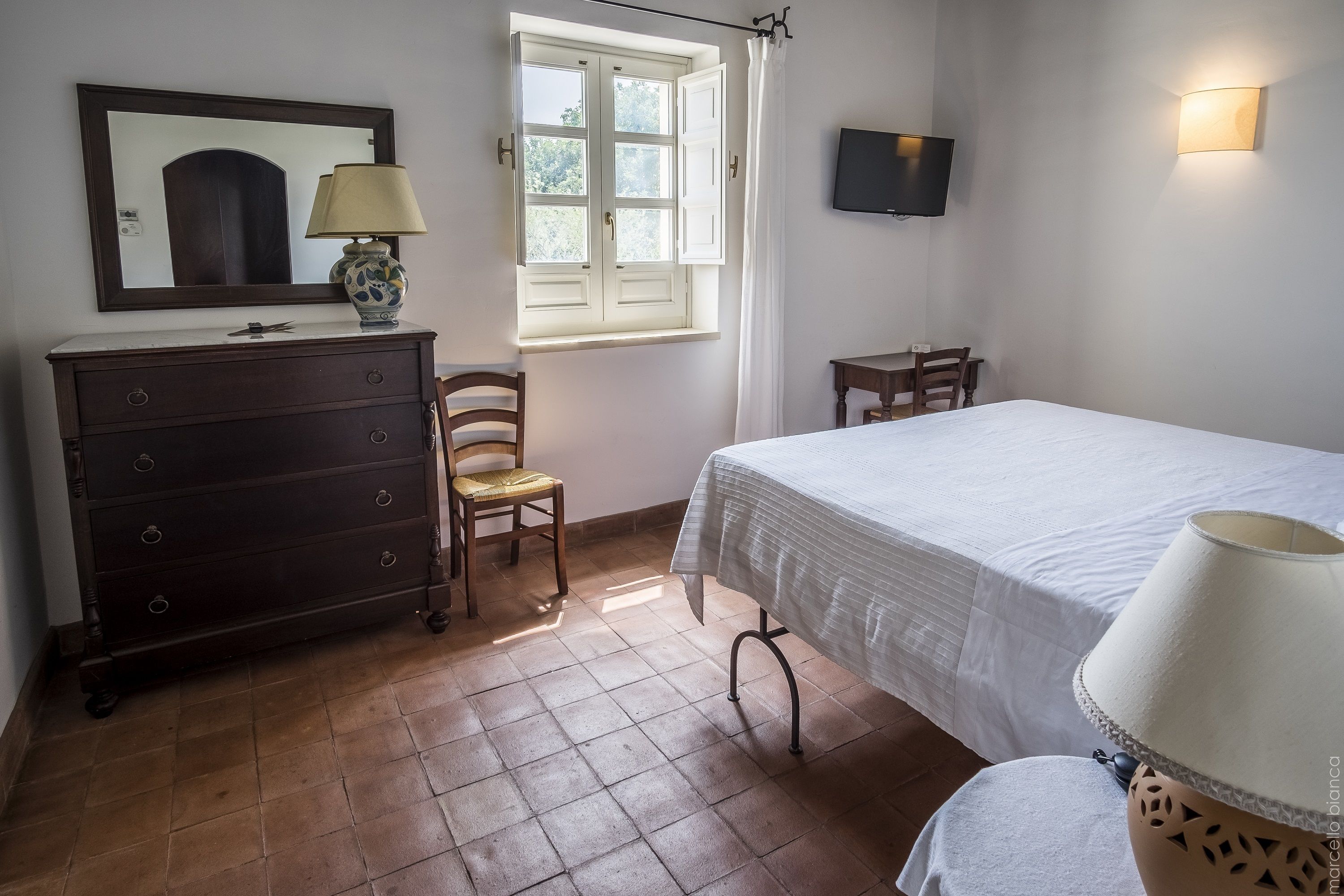 Une chambre à coucher comprenant un lit, une commode, une chaise, une fenêtre avec des rideaux, un téléviseur fixé au mur et une lampe sur la commode.