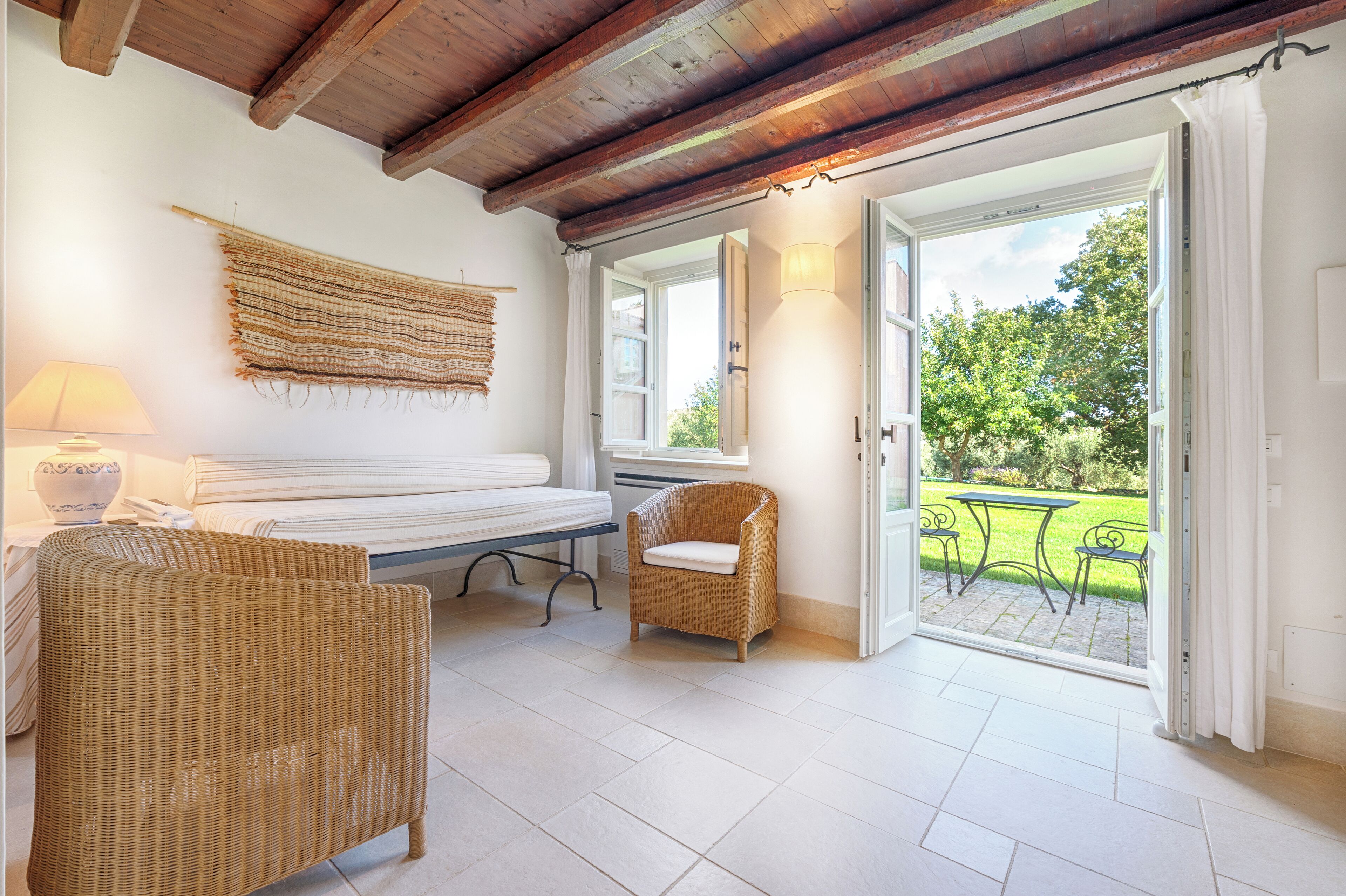 Un dormitorio con una cama, una silla de mimbre, una lámpara y vista a un jardín desde una puerta abierta.
