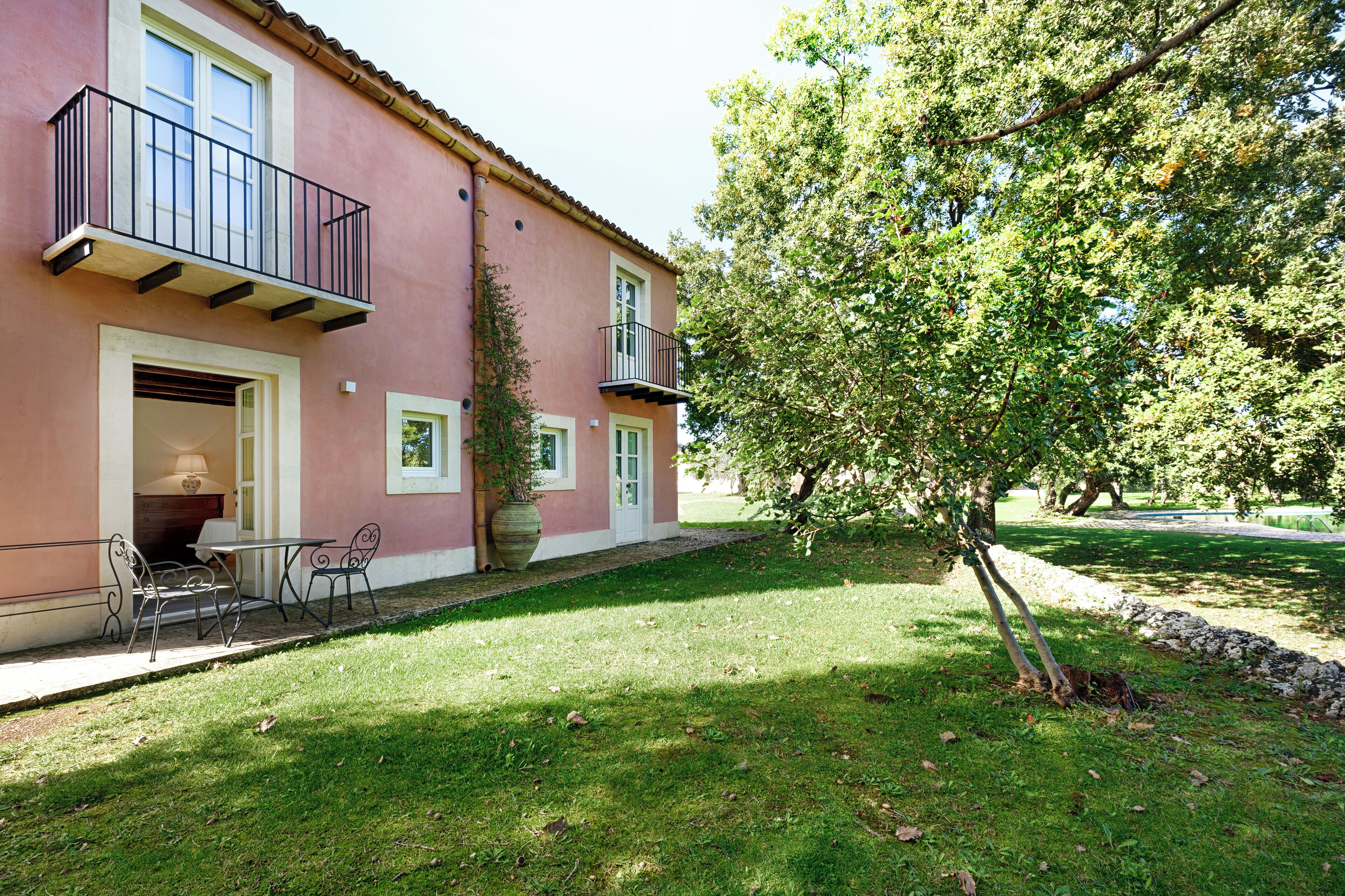 Un edificio rosa con un balcón, un pequeño patio con sillas y un árbol en el jardín.