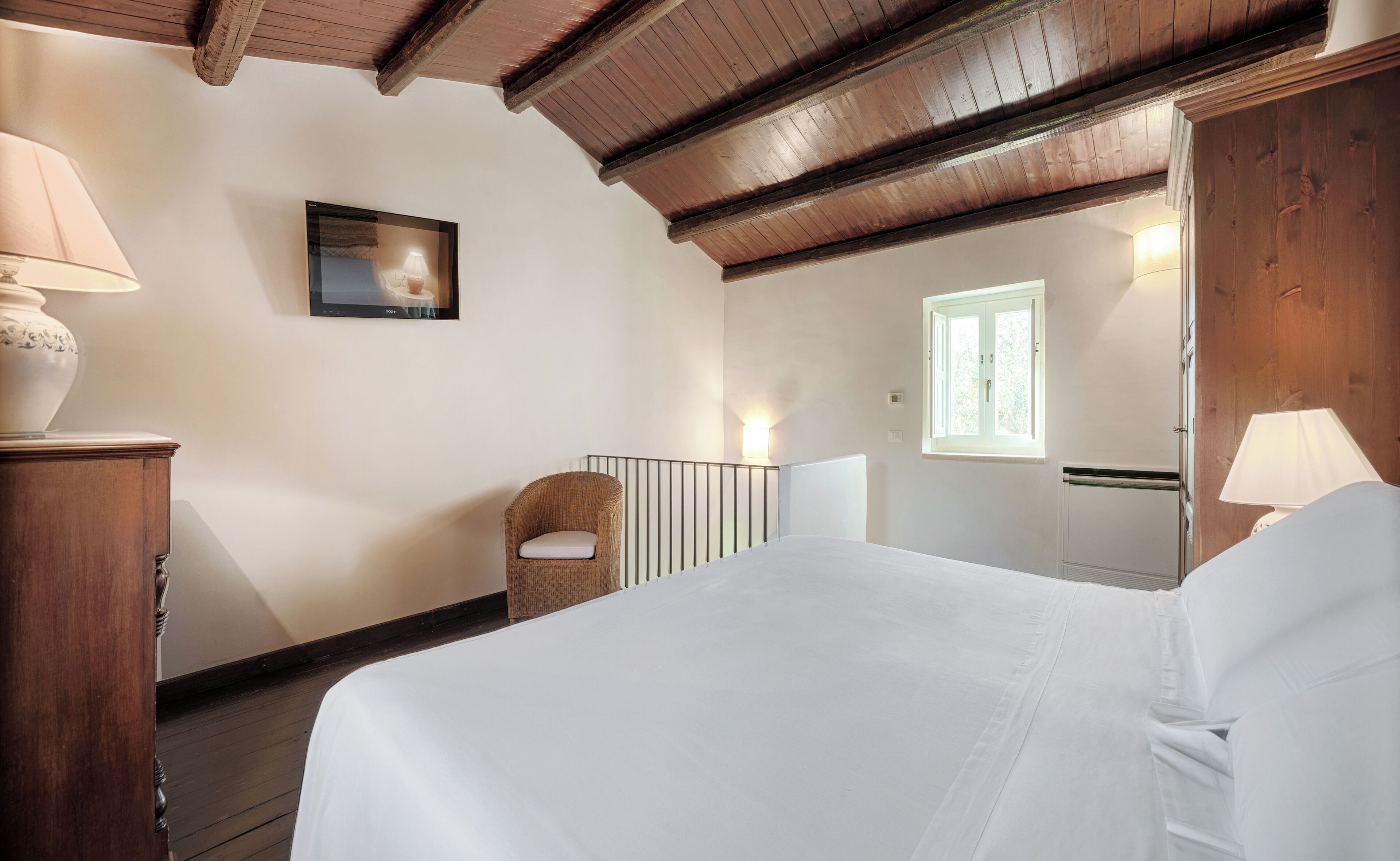 Un dormitorio con techo de madera, una cama, una silla, una mesita de noche, una lámpara, un cuadro y una ventana.