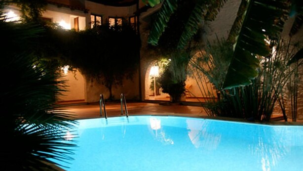Une piscine entourée d’une végétation luxuriante et un bâtiment dont les fenêtres sont illuminées.