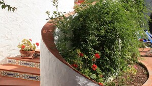 Un bac à fleurs blanc et incurvé, orné de diverses plantes et fleurs, un escalier carrelé et un mur blanc surmonté d’un toit en tuiles.