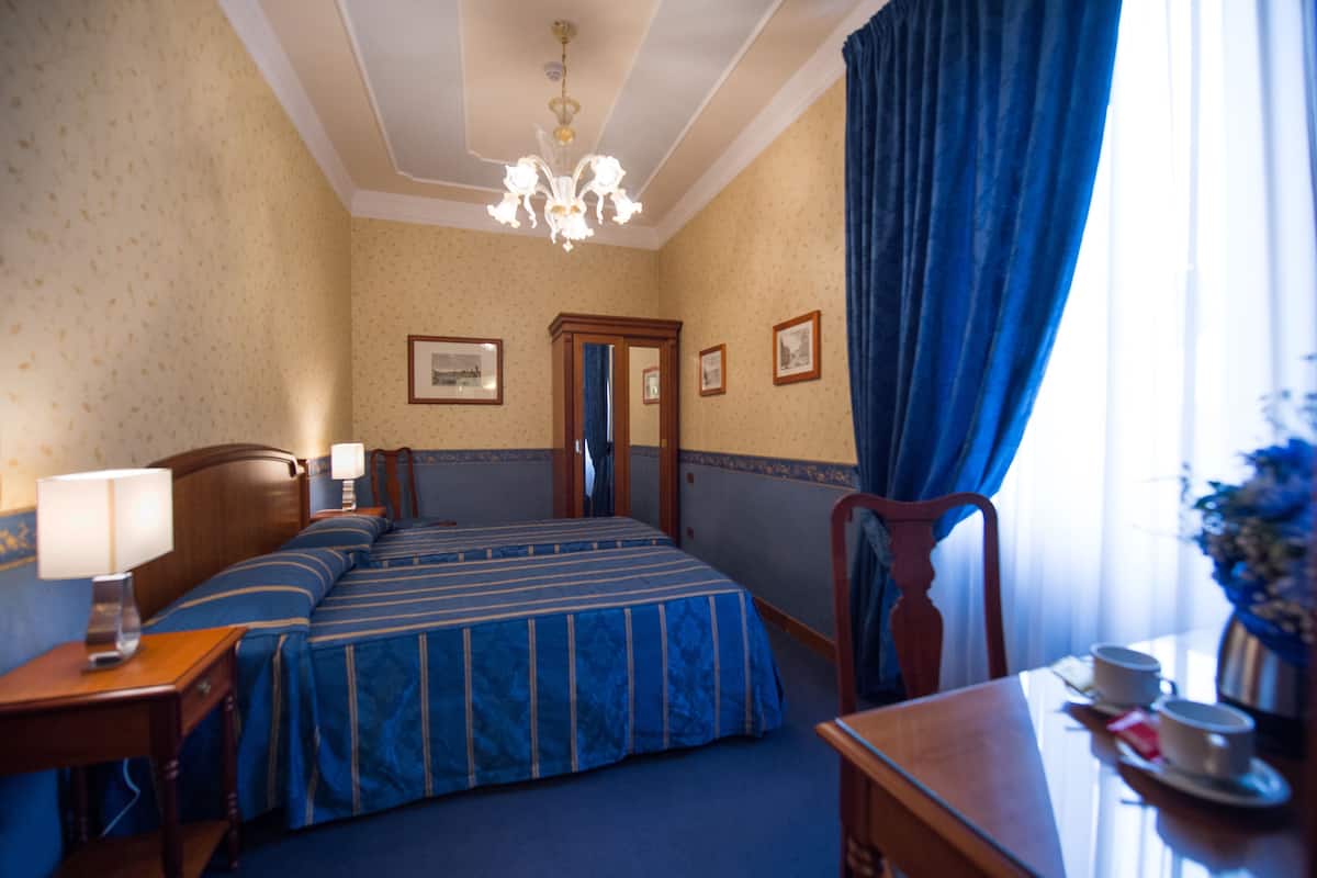 Une chambre d’hôtel avec un lit double, une table de chevet en bois avec une lampe, un bureau avec une tasse et une soucoupe, et une grande fenêtre avec des rideaux bleus.