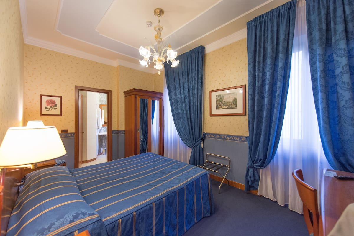 Une chambre d’hôtel avec un lit, une chaise, une table, une lampe et un lustre.