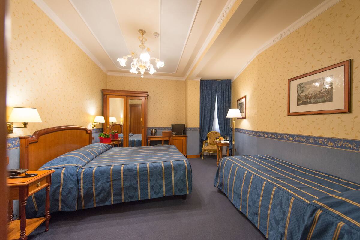 Une chambre d’hôtel avec deux lits, un bureau, une chaise, un miroir, un lustre et un tableau encadré au mur.