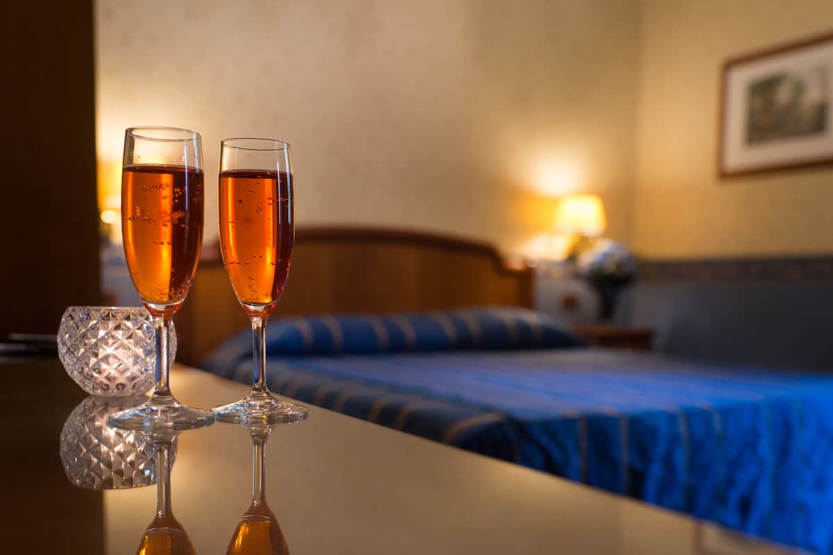 Deux coupes à champagne posées sur une surface réfléchissante, un lit recouvert d’une couverture bleue et une table de chevet avec une lampe, dans une chambre d’hôtel.