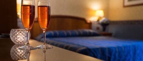 Dos copas de champán sobre una superficie reflectante, una cama con cubrecama azul y una mesita de noche con lámpara en una habitación de hotel.