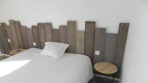 A bedroom with a wooden headboard, a white bedspread, a white pillow, and two round wooden nightstands with black wireframe bases. - Hotel Les Provinciales (Arpajon-sur-Cere)