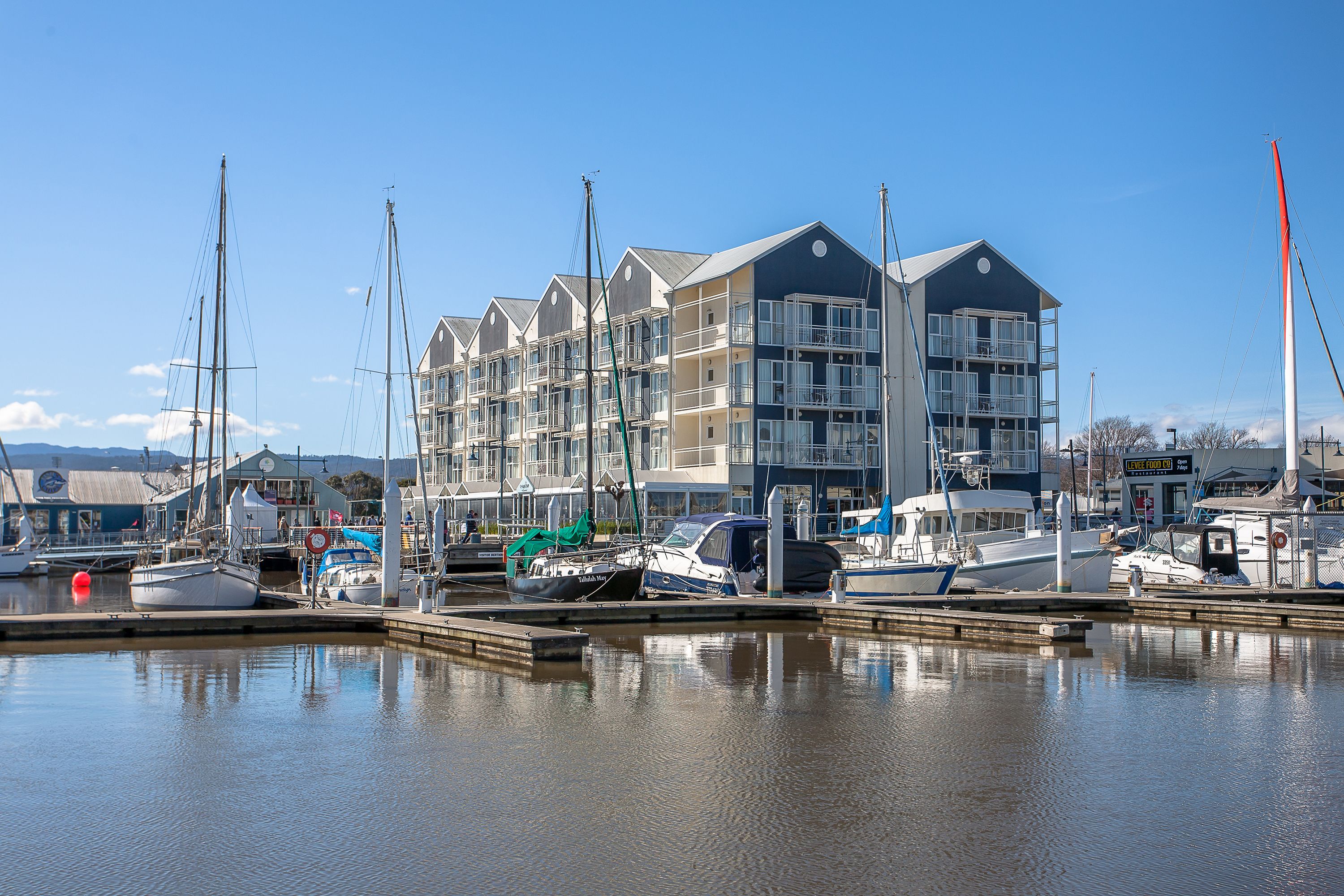 A marina with several boats docked, a multi-story building with balconies, and a clear blue sky.