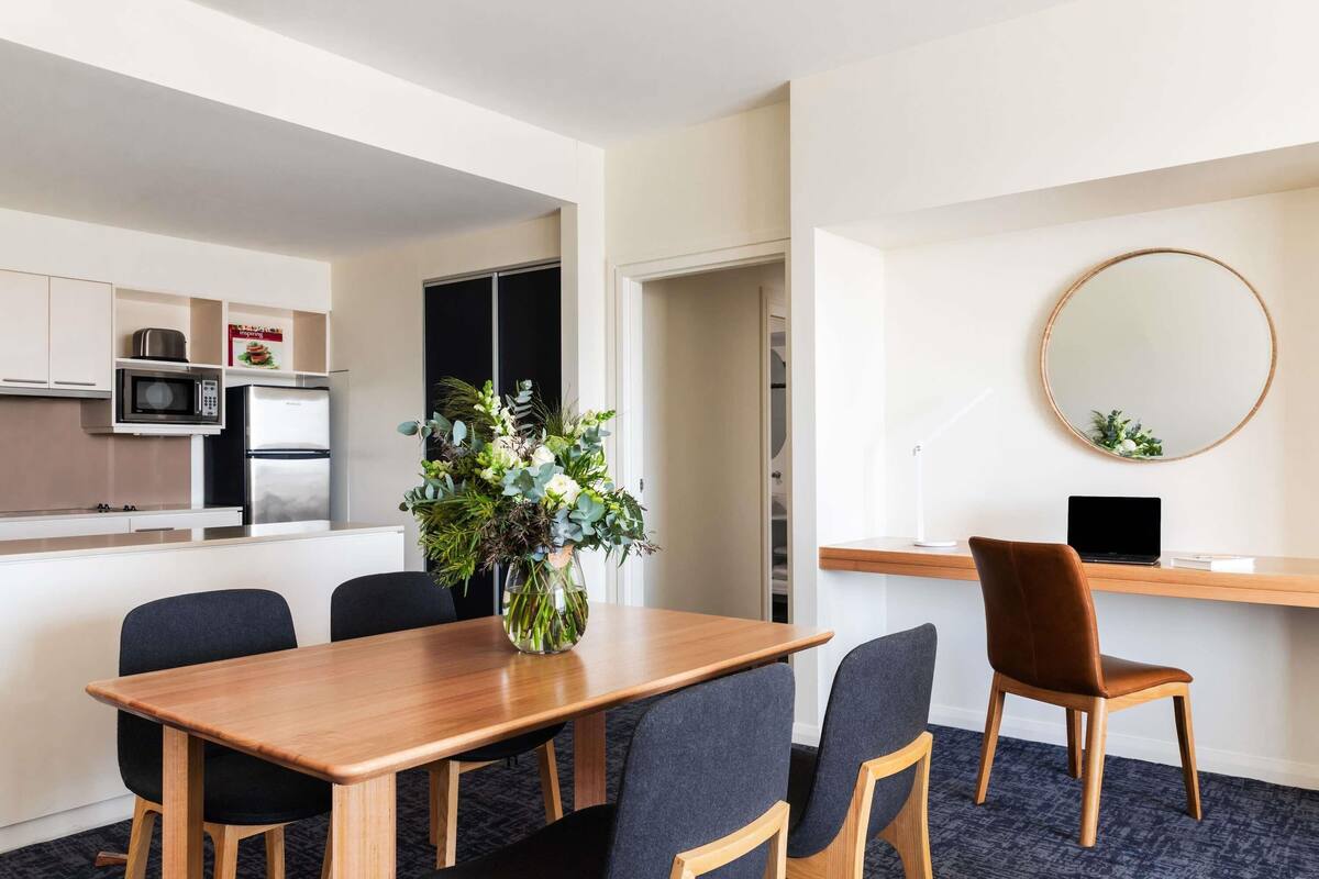 A modern hotel room with a wooden dining table, four chairs, a laptop on a desk, a mirror, and a kitchen area with appliances.
