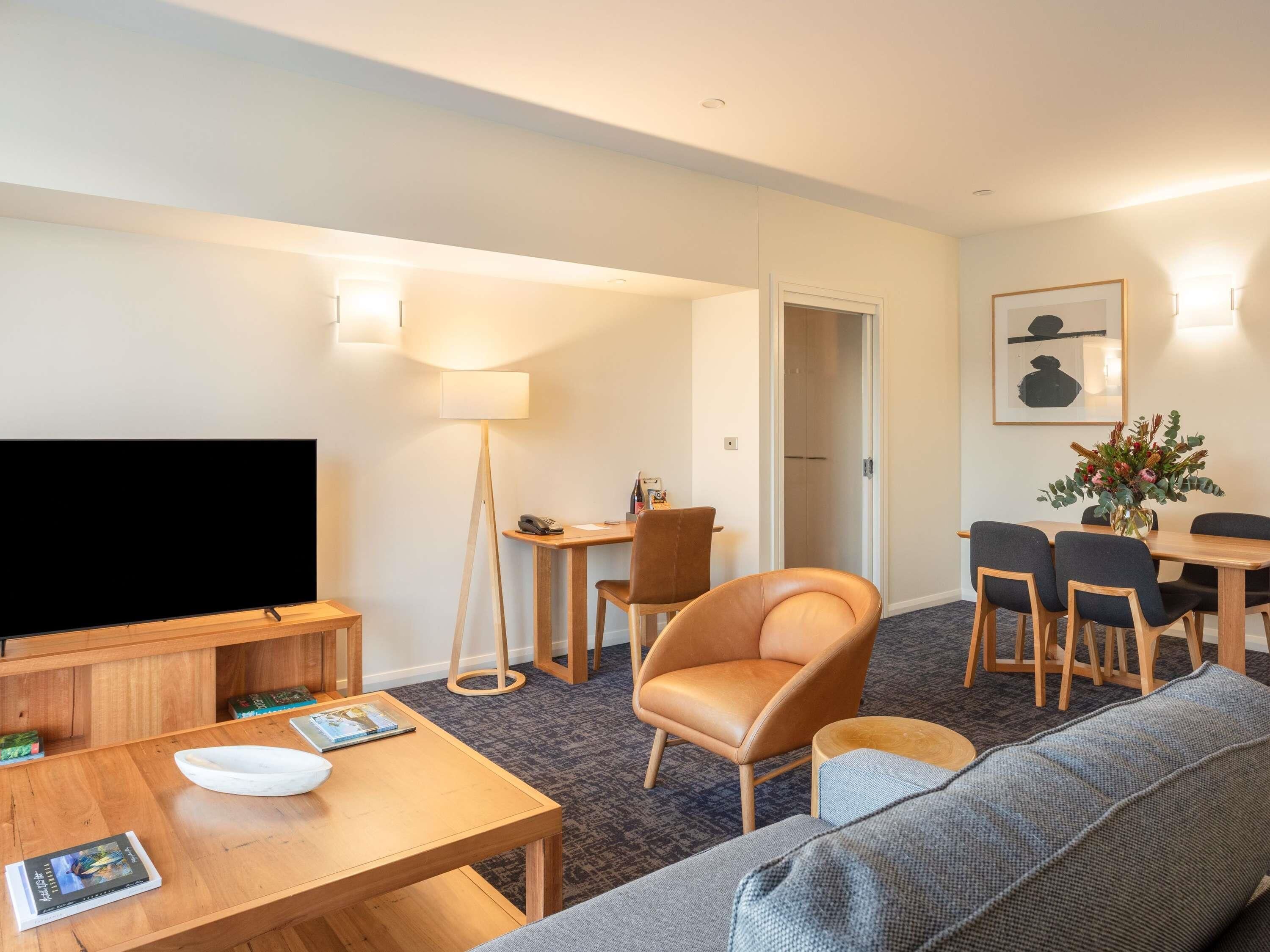 A hotel room with a flat-screen TV, wooden furniture, a sofa, a dining table with chairs, a lamp, and a framed picture on the wall.