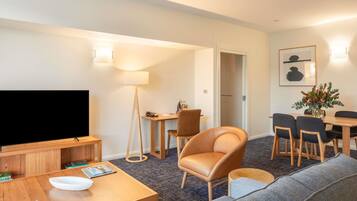 A hotel room with a flat-screen TV, wooden furniture, a sofa, a dining table with chairs, a lamp, and a framed picture on the wall.