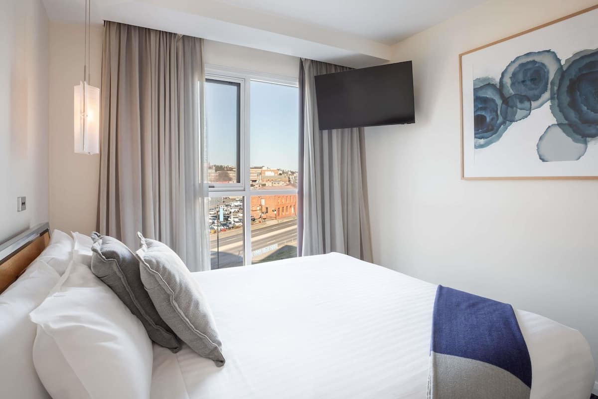 A hotel room with a large bed, a flat-screen TV, a painting, and a view of the city through the window.